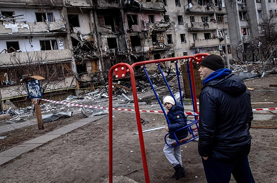 A boy plays on a swing in front of a damaged residential building hit by an early morning strike in Feb. 2022 in Kyiv, Ukraine / www.president.gov.ua